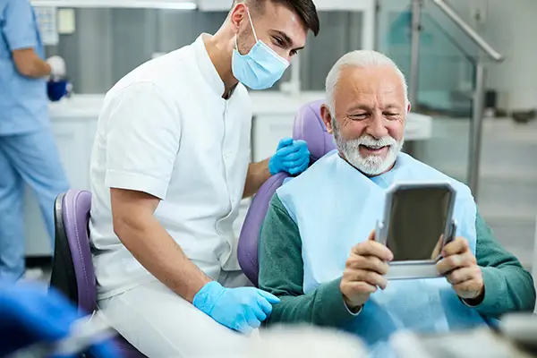 Smiling senior patient with dentures looking at himself in a hand mirror